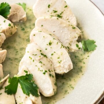 Round plate with slices of Poached Chicken Breast, sitting on a lemon parsley sauce with some pieces of parsley as a garnish.