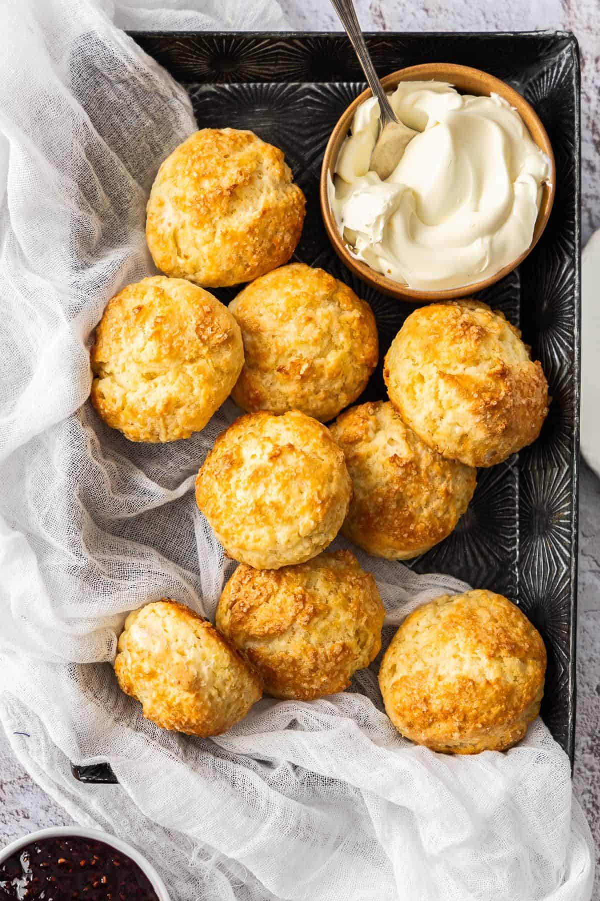 Black tray with a white cloth, full of Buttermilk Scones, with a small dish of jam and cream in the corners of the trays.