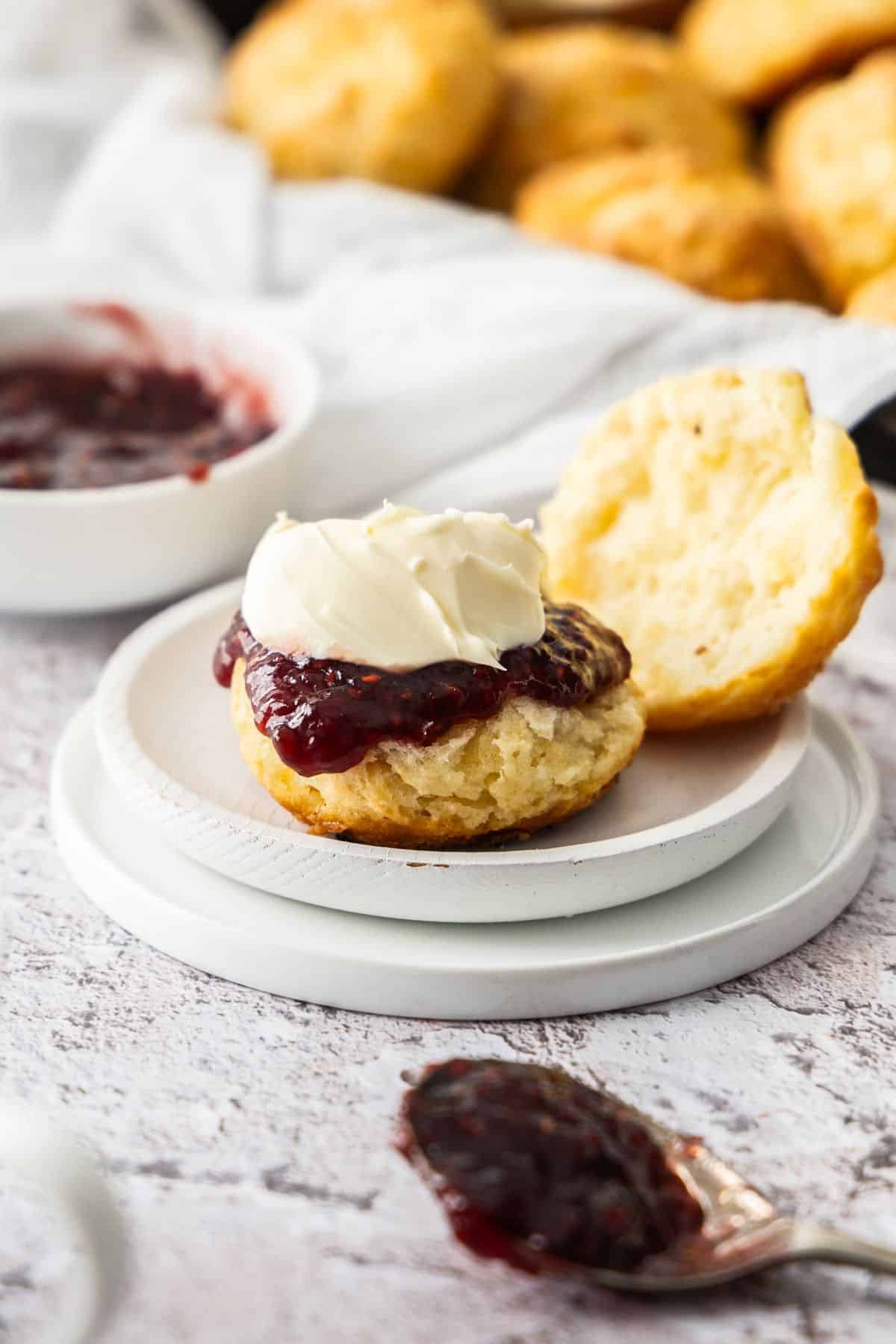 A scone, cut in half, sitting on a small round white plate, topped with raspberry jam and whipped cream.