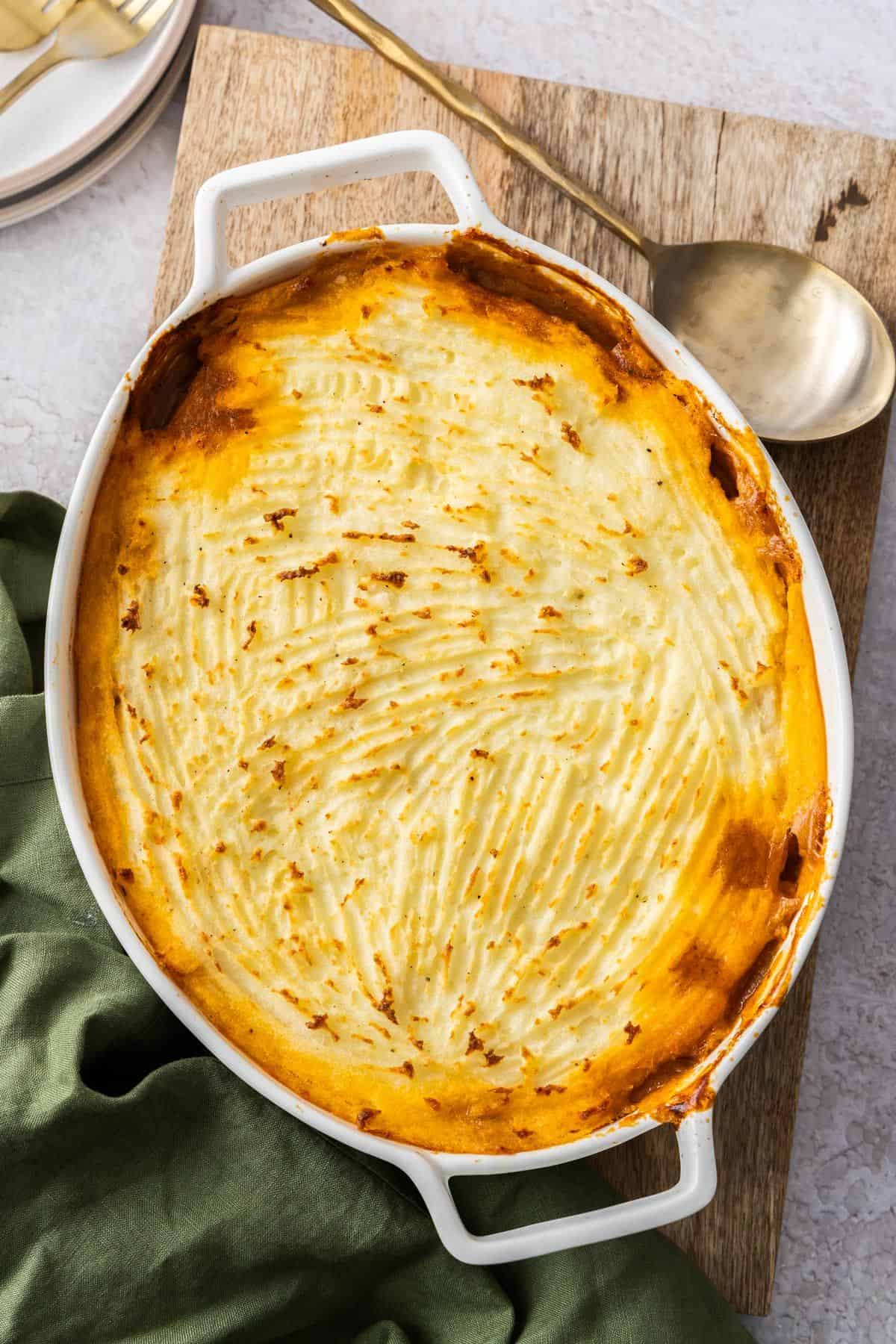 White oval dish of Cottage Pie, sitting on a green cloth and wooden board, with a gold spoon on the edge of the board.