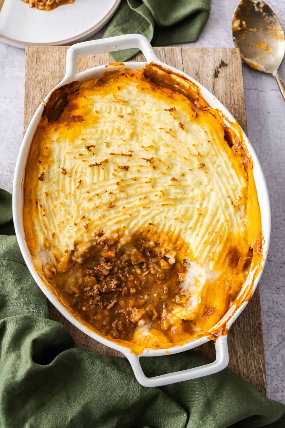 White oval dish of Cottage Pie, sitting on a green cloth and wooden board, with a serving having been removed from the dish.