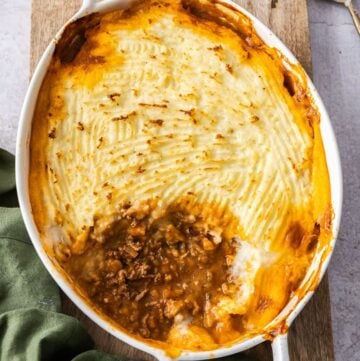 White oval dish of Cottage Pie, sitting on a green cloth and wooden board, with a serving having been removed from the dish.