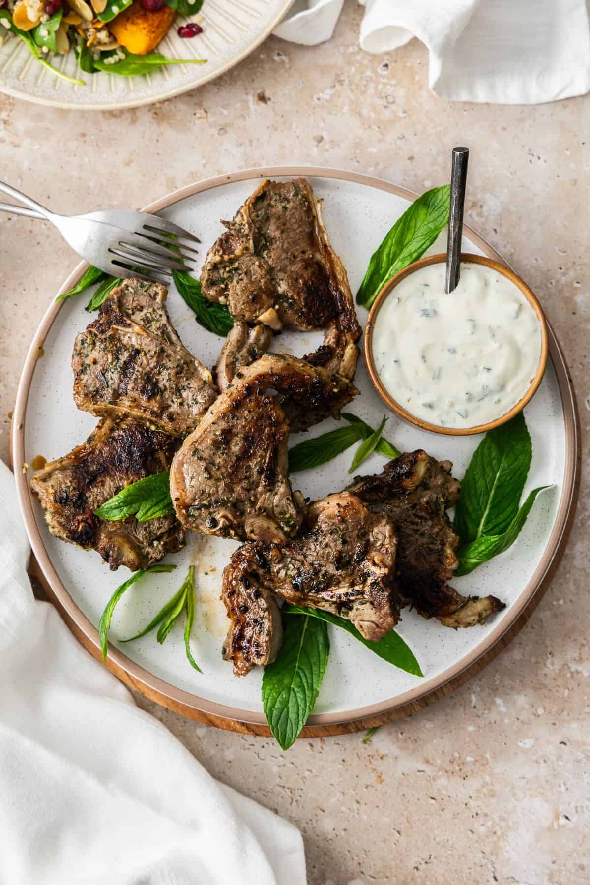 Round white plate with cooked Marinated Lamb Loin Chops, with a small bowl of yoghurt sauce on the side, and white cloth.