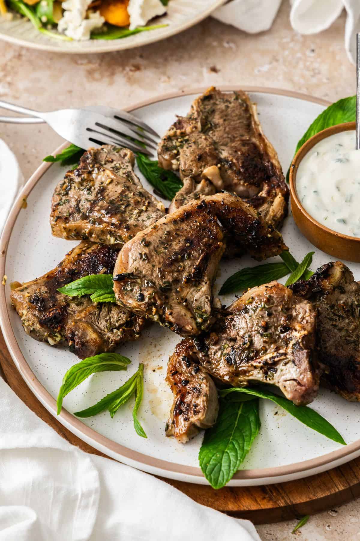 Round white plate with cooked Marinated Lamb Loin Chops, with a small bowl of yoghurt sauce on the side, and white cloth.