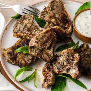 Round white plate with cooked Marinated Lamb Loin Chops, with a small bowl of yoghurt sauce on the side, and white cloth.