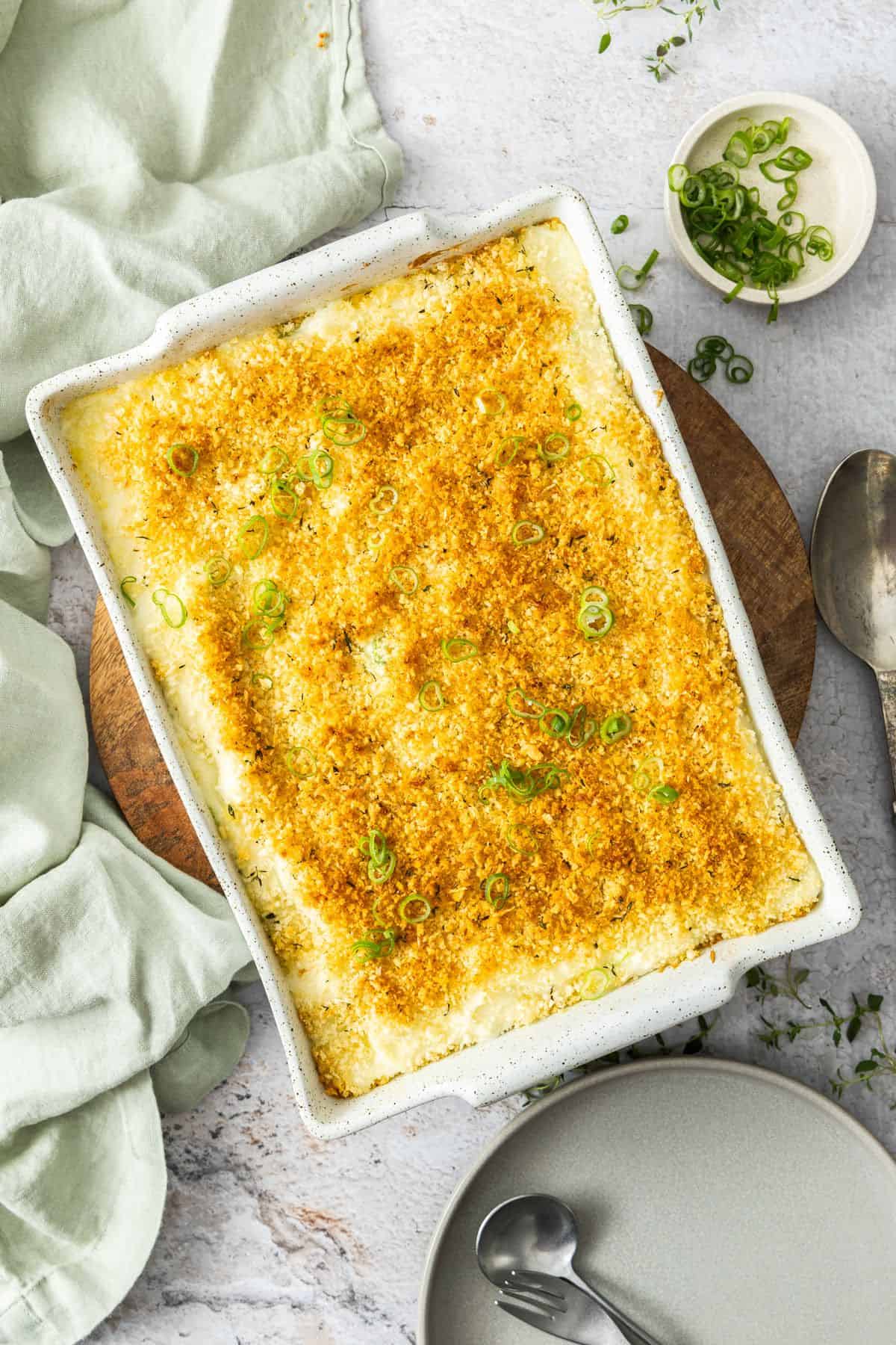 Rectangular dish of Baked Mashed Potatoes, sitting on a round wooden board with a green cloth on the edge.