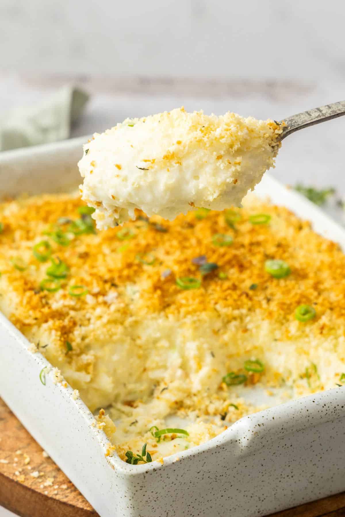 Rectangular dish of Baked Mashed Potatoes with a spoon lifting up a serve from the dish.