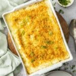 Rectangular dish of Baked Mashed Potatoes, sitting on a round wooden board with a green cloth on the edge.