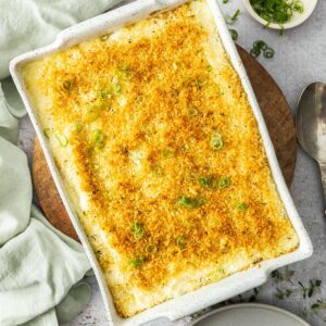 Rectangular dish of Baked Mashed Potatoes, sitting on a round wooden board with a green cloth on the edge.