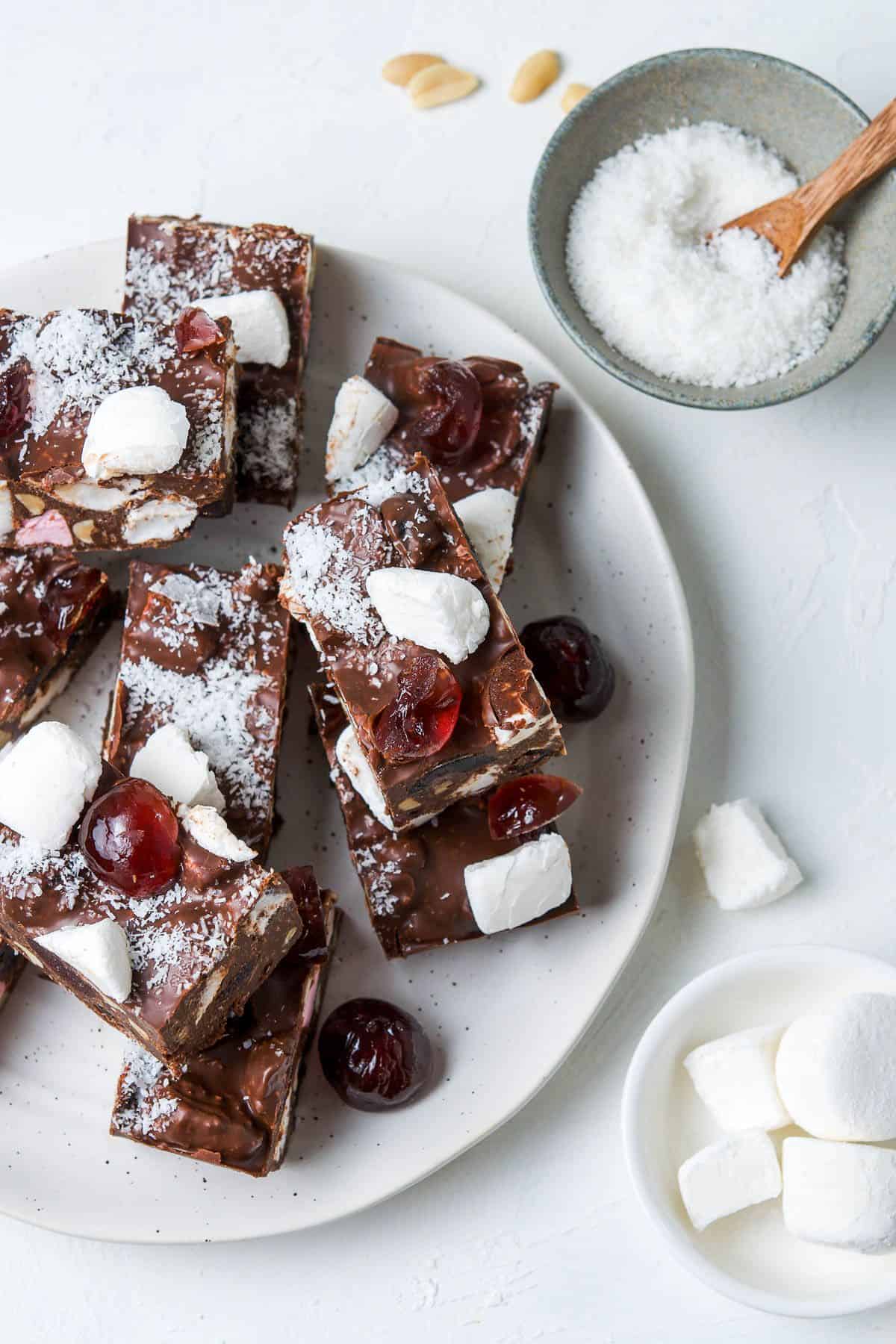 Round white plate with cut pieces of Rocky Road, sitting on a white bench top, with small dishes of coconut and marshmallows.