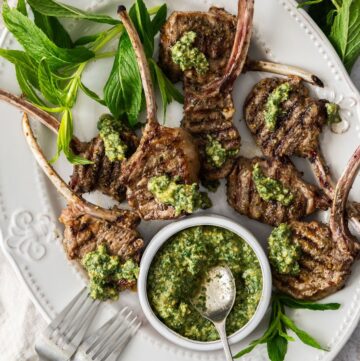 Oval white plate with Grilled Lamb Chops, some fresh mint leaves, two forks and a small dish of Mint Pesto.