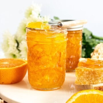 Two jars of Orange Marmalade, one with a spoon on it, sitting on a round white plate with some slices of bread.