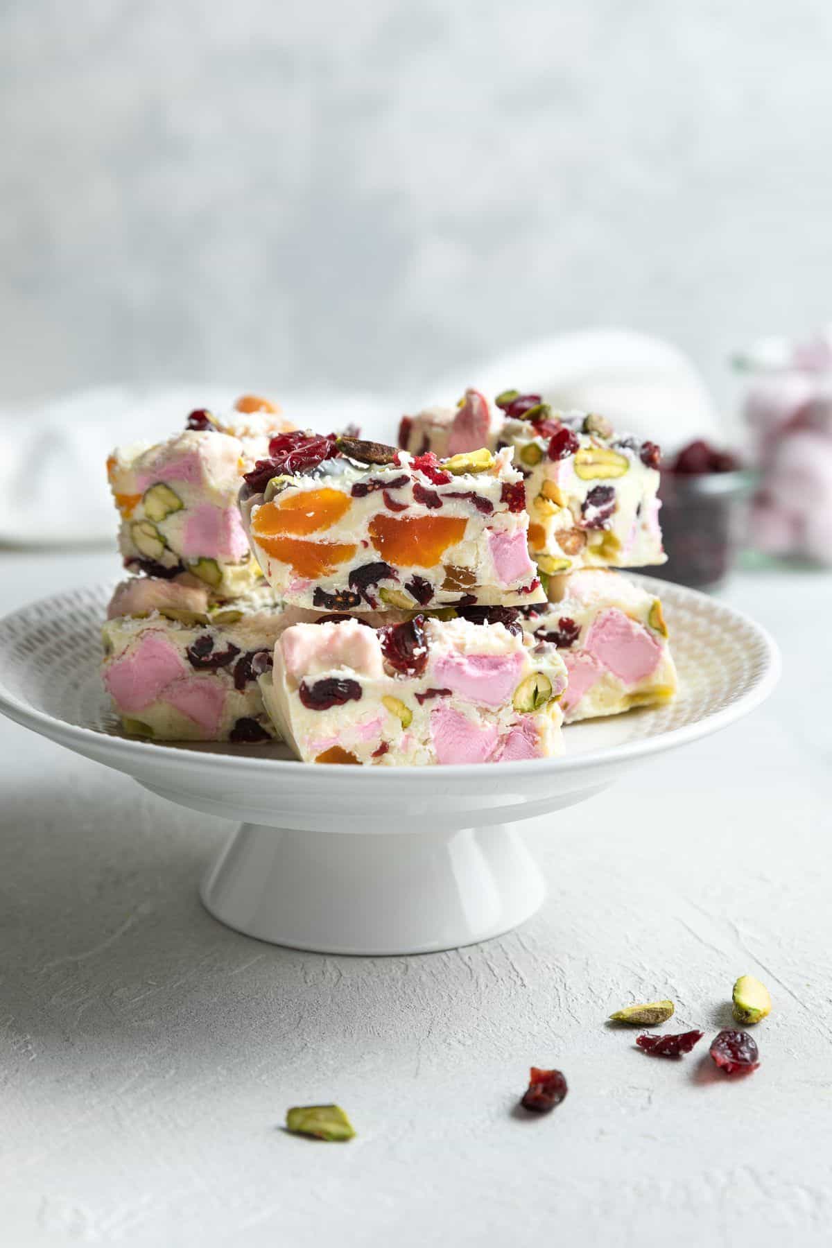 Round tall cake stand with cut pieces of Rocky Road, sitting on a white bench top.