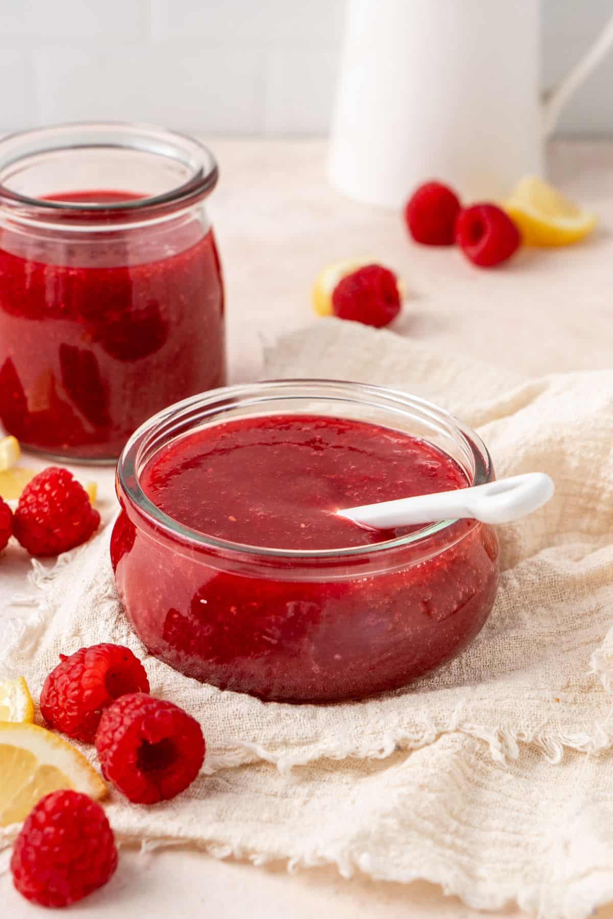 Round glass dish of Raspberry Sauce sitting on a white cloth, with a spoon resting in the dish.