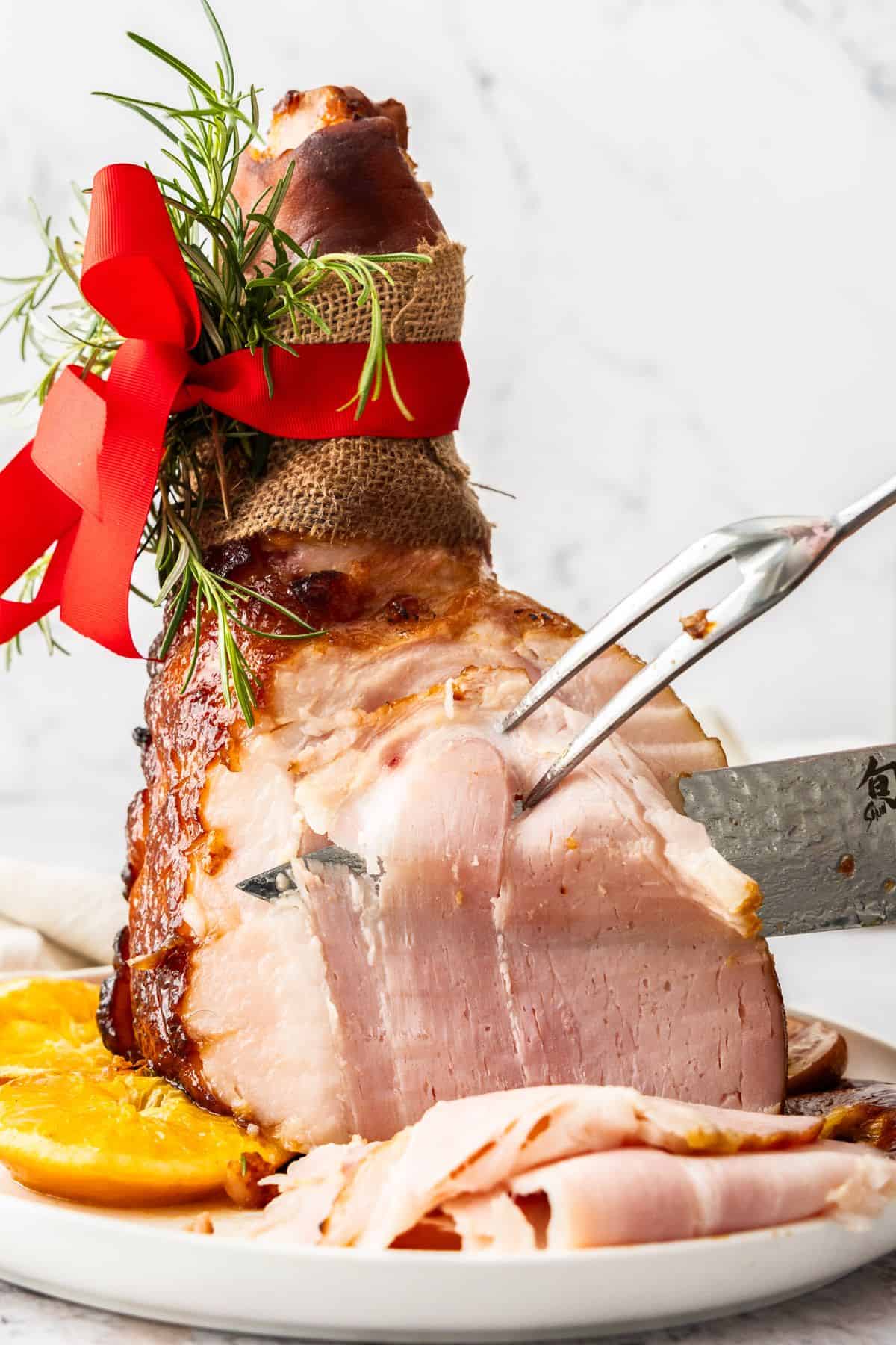 Maple Glazed Ham, sitting on a round white plate, with a fork and knife assisting with carving some slices.