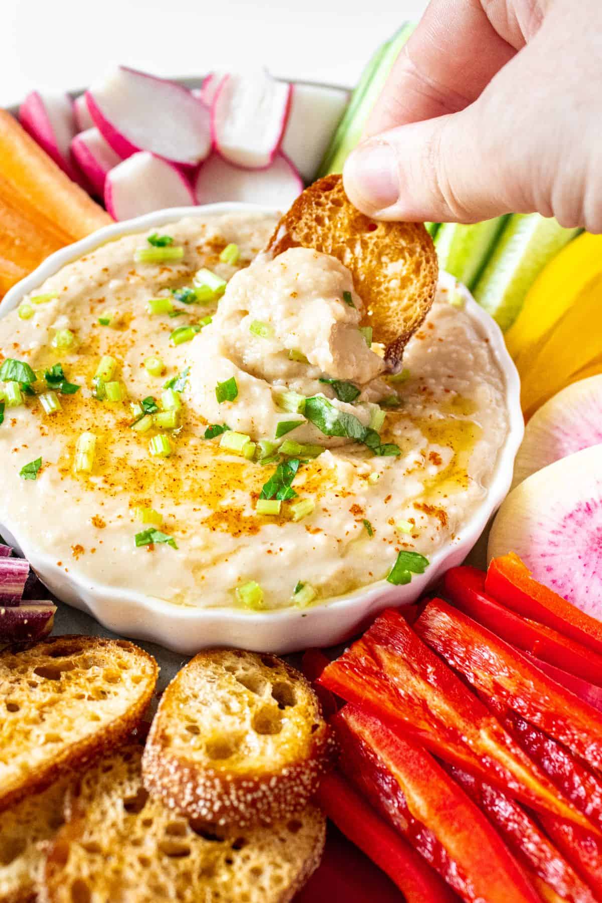 Dip platter featuring Butter Bean Dip and a hand scooping up some dip on a toast.