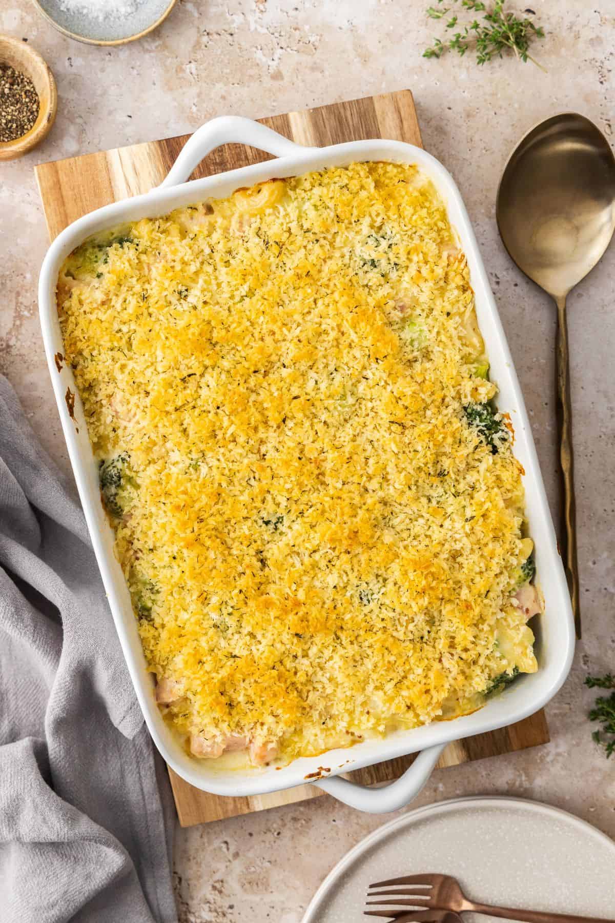 White rectangular dish of Creamy Ham Pasta Bake, sitting on a wooden board with a serving spoon on the edge.