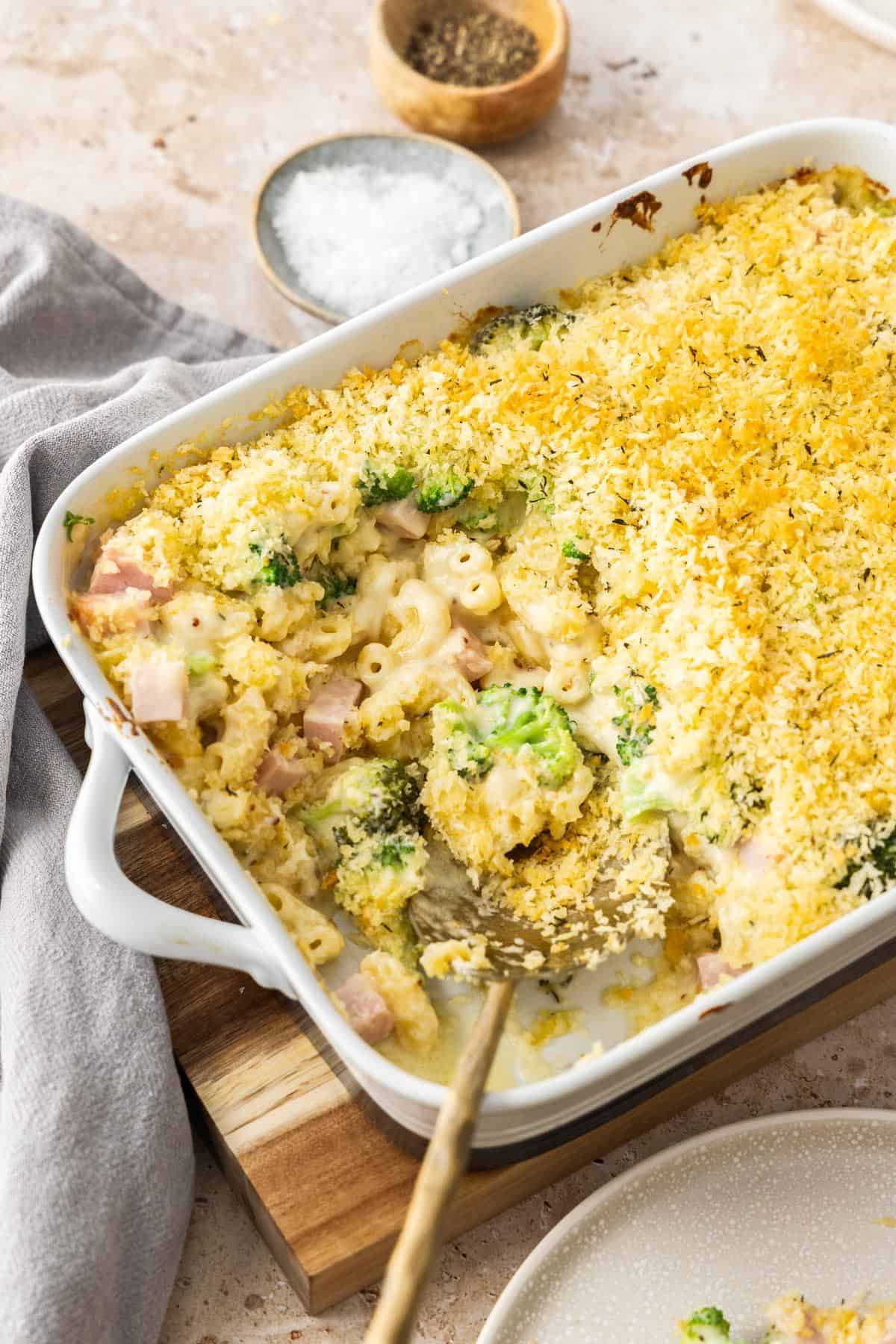 White rectangular dish of Creamy Ham Pasta Bake, sitting on a wooden board with a serve having been removed.