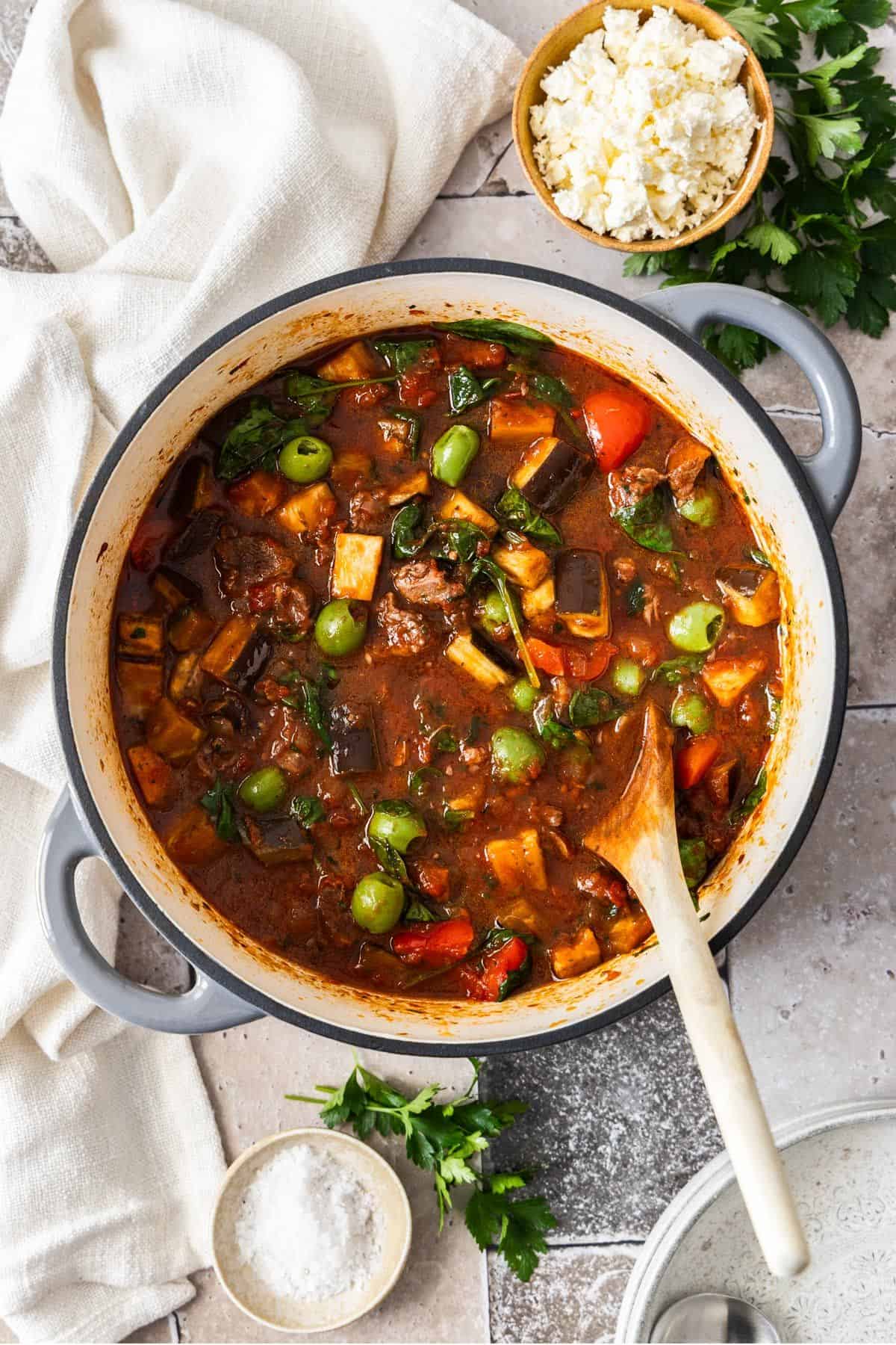 Mediterranean Lamb Stew, in a large grey saucepan sitting on a white cloth, with a wooden spoon resting in the pan.