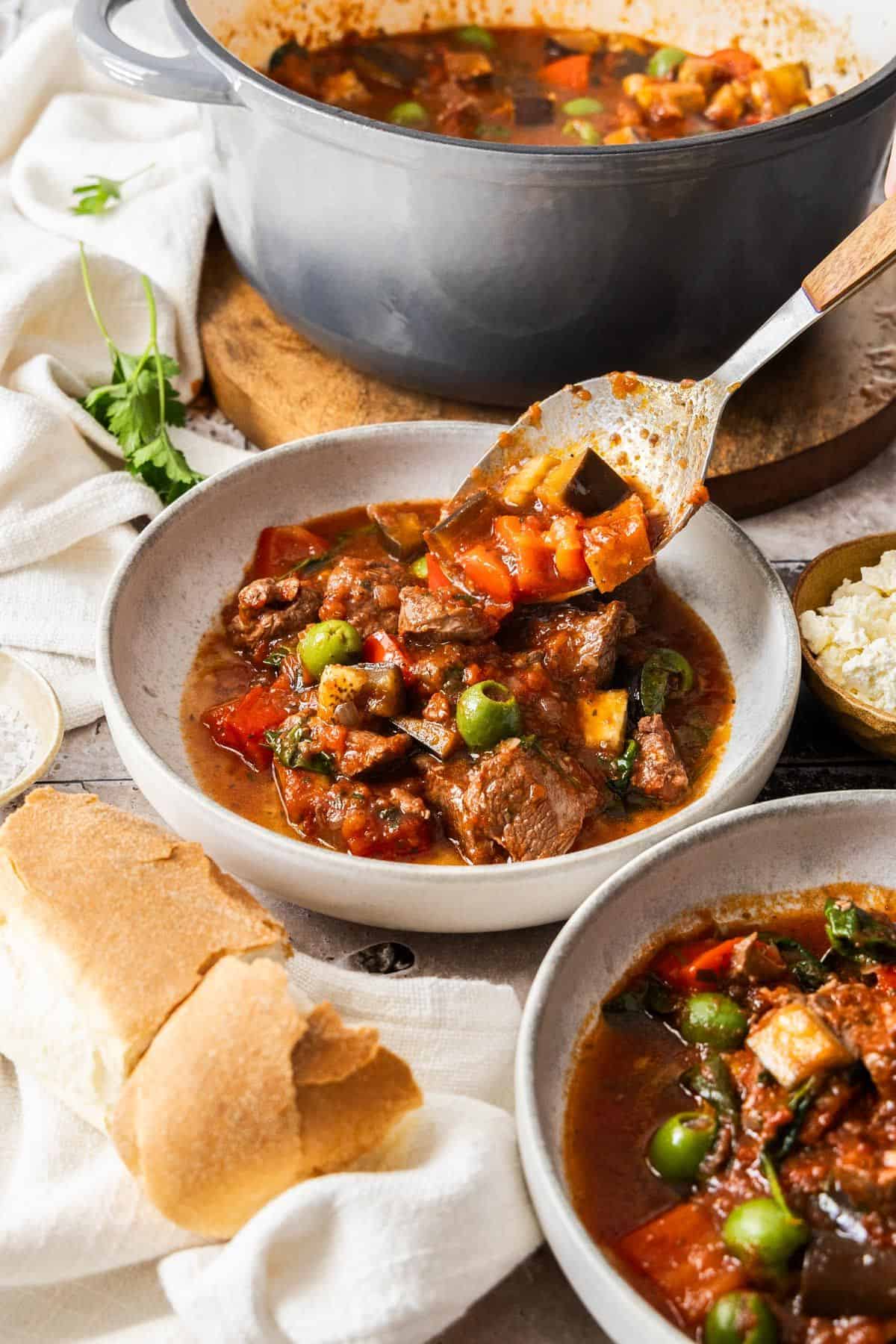 Spooning the Lamb Stew into a round grey bowl, with another bowl of stew on the side, and pot of stew sitting behind.