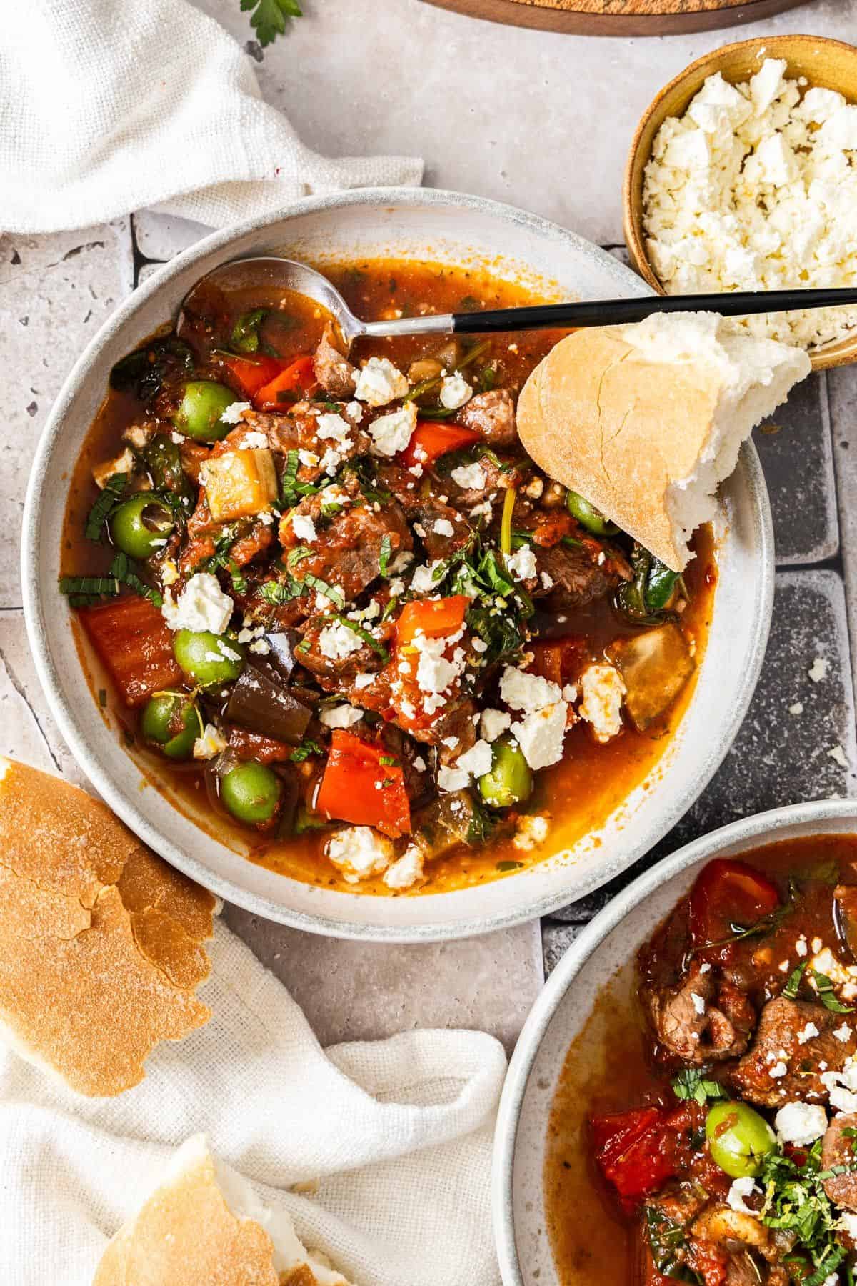 The Lamb Stew in a round grey bowl with a spoon, topped with feta and herbs and a piece of bread on the edge.