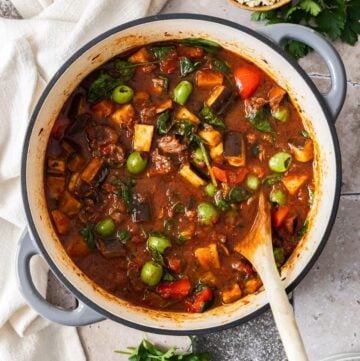 Mediterranean Lamb Stew, in a large grey saucepan sitting on a white cloth, with a wooden spoon resting in the pan.