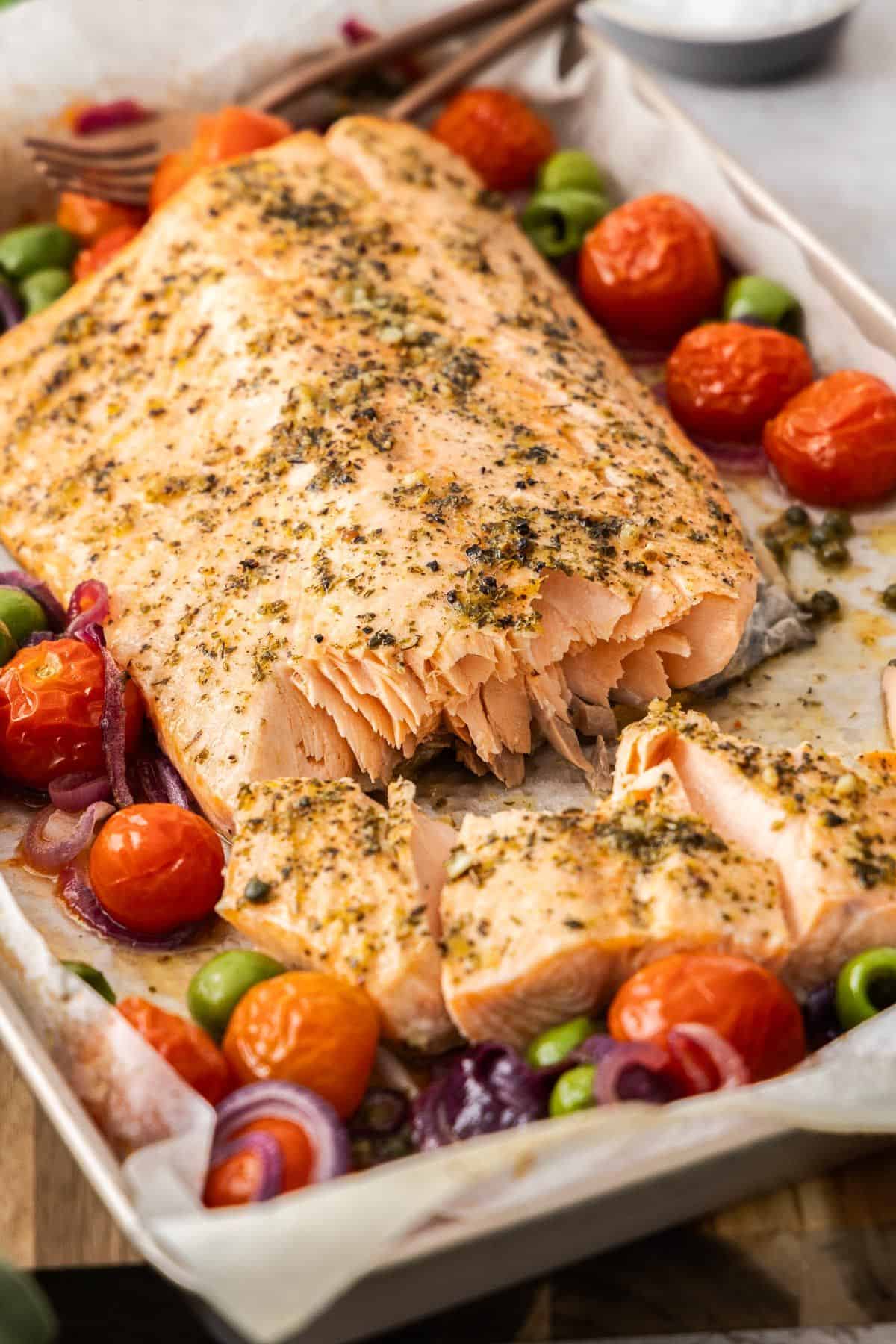 Sheet pan with side of Mediterranean Salmon, broken up slightly, and vegetables around the edge, with serving utensils.