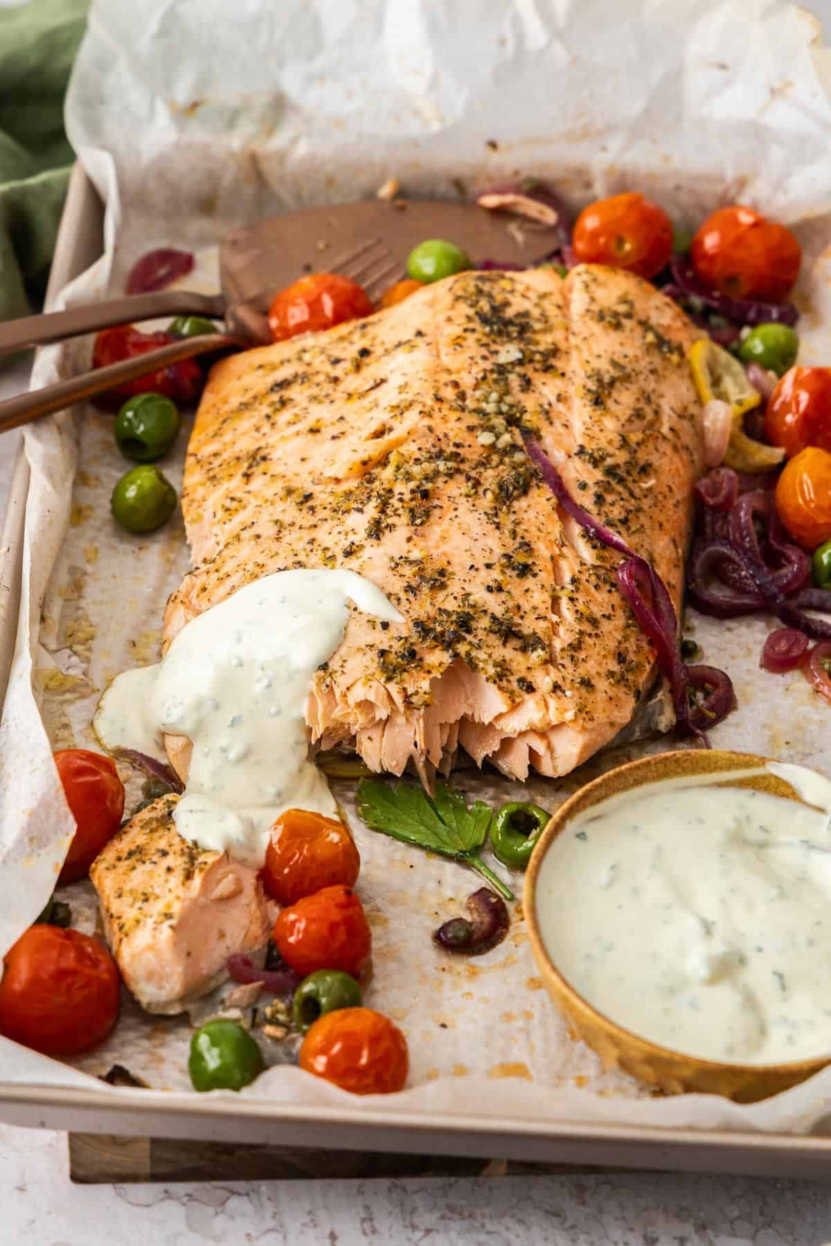 Sheet pan with side of Mediterranean Salmon, broken up slightly, and vegetables around the edge, with serving utensils.