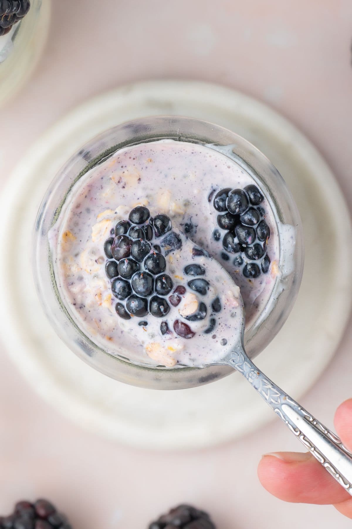 Jar of Blackberry Overnight Oats, sitting on a small white saucer, with a spoon scooping up some of the mixture.