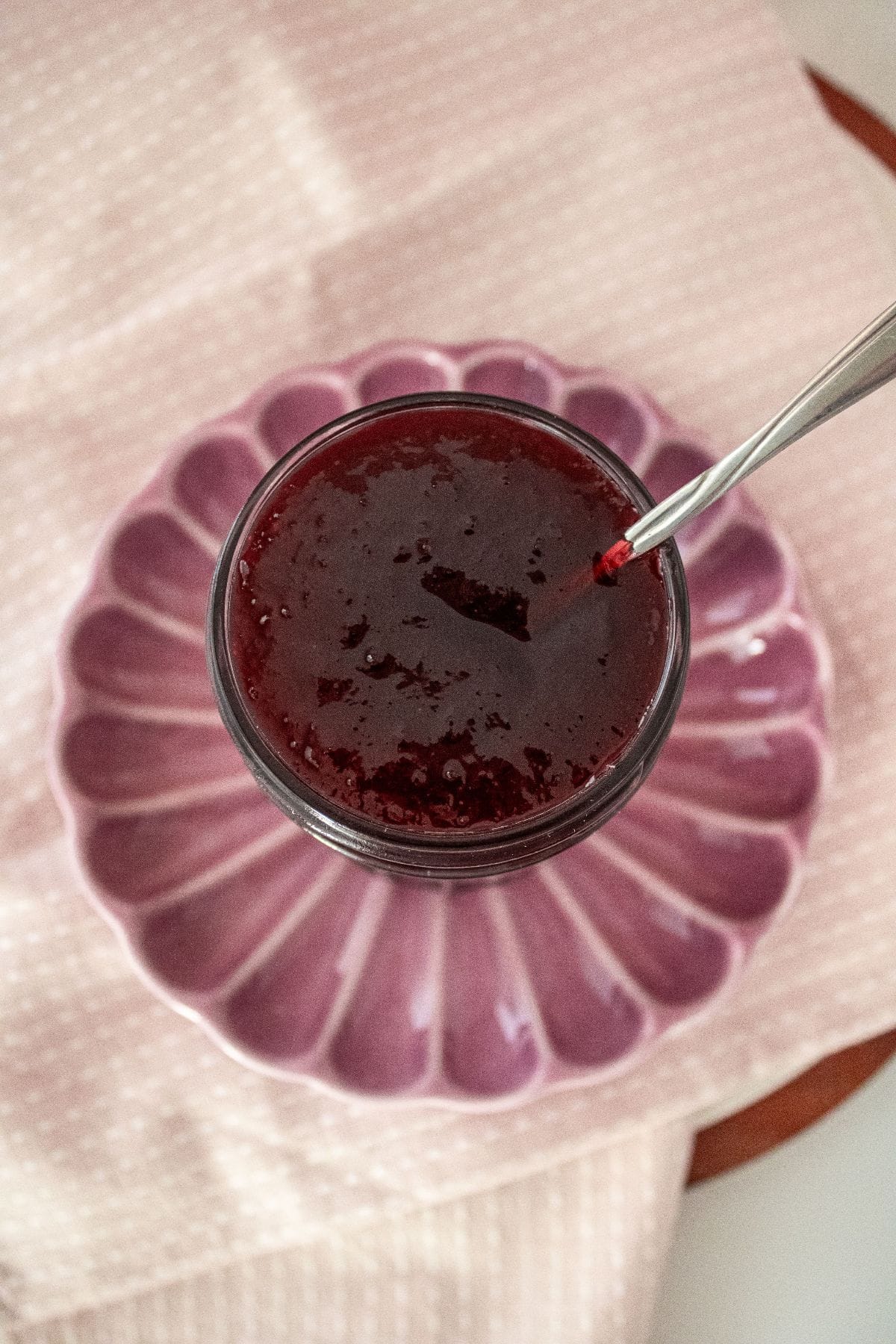 Open jar of Cherry Jam on a round pink plate, with a spoon sitting in jar.