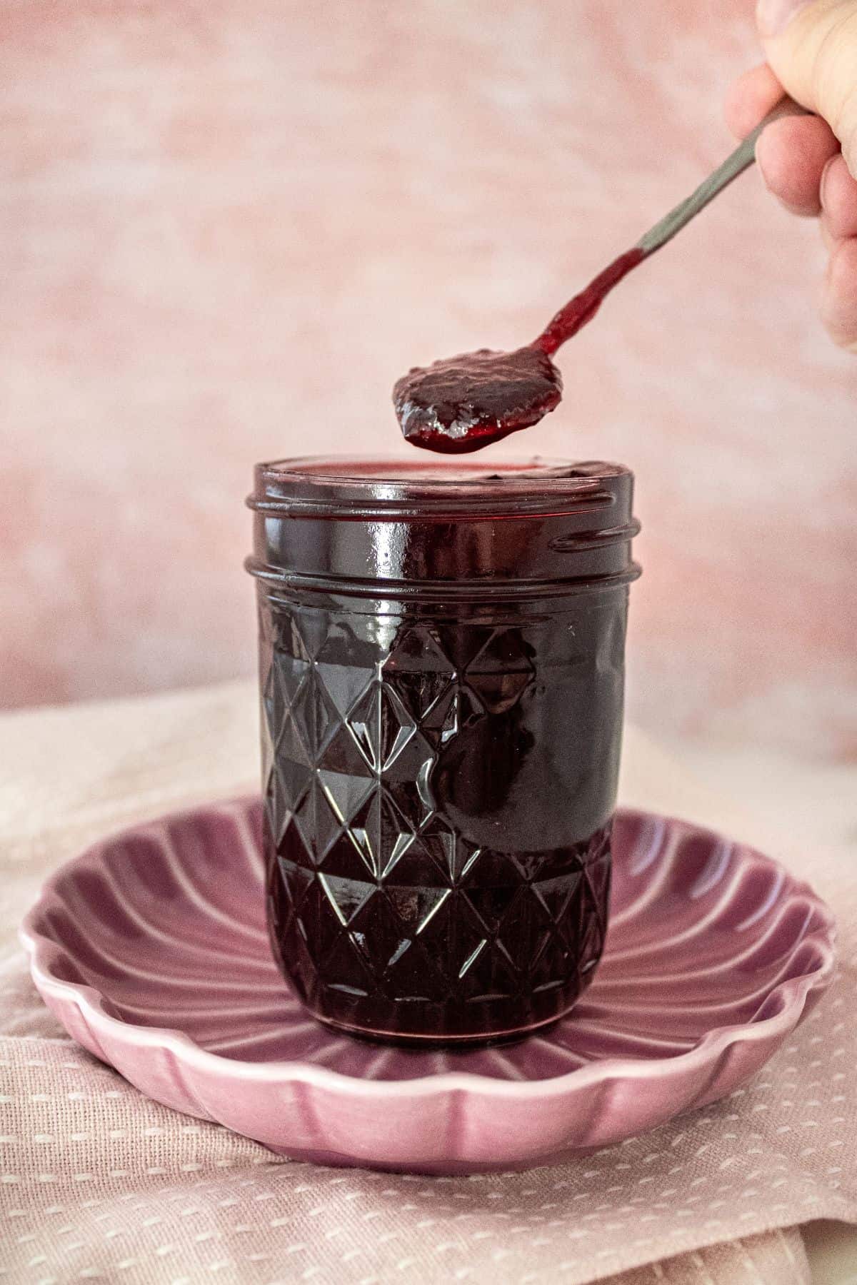 Open jar of Cherry Jam on a round pink plate, with a spoon scooping up some jam.