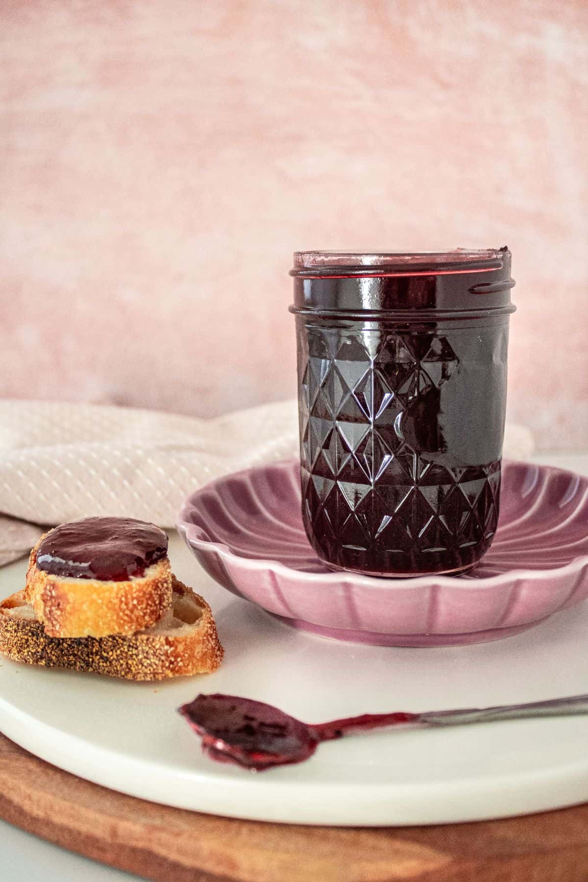 Open jar of Cherry Jam on a round pink plate, with a spoon resting on the edge and some pieces of bread with jam.