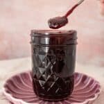 Open jar of Cherry Jam on a round pink plate, with a spoon scooping up some jam.