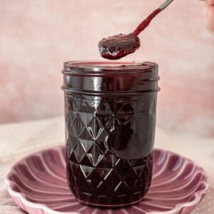 Open jar of Cherry Jam on a round pink plate, with a spoon scooping up some jam.