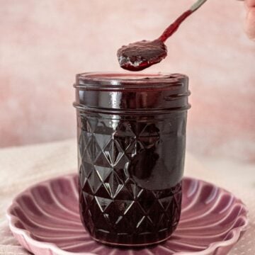 Open jar of Cherry Jam on a round pink plate, with a spoon scooping up some jam.