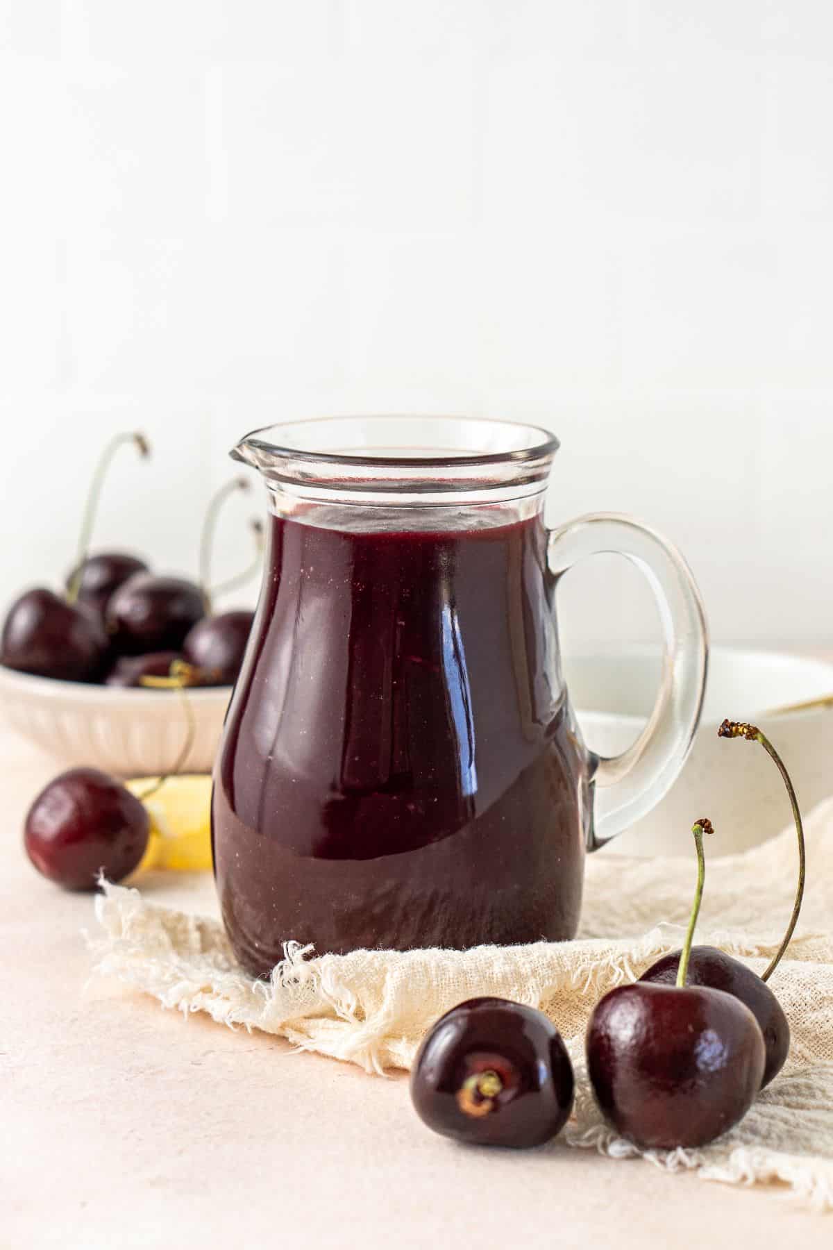 Jug of Cherry Sauce, on a cream cloth with some fresh cherries around the edge.