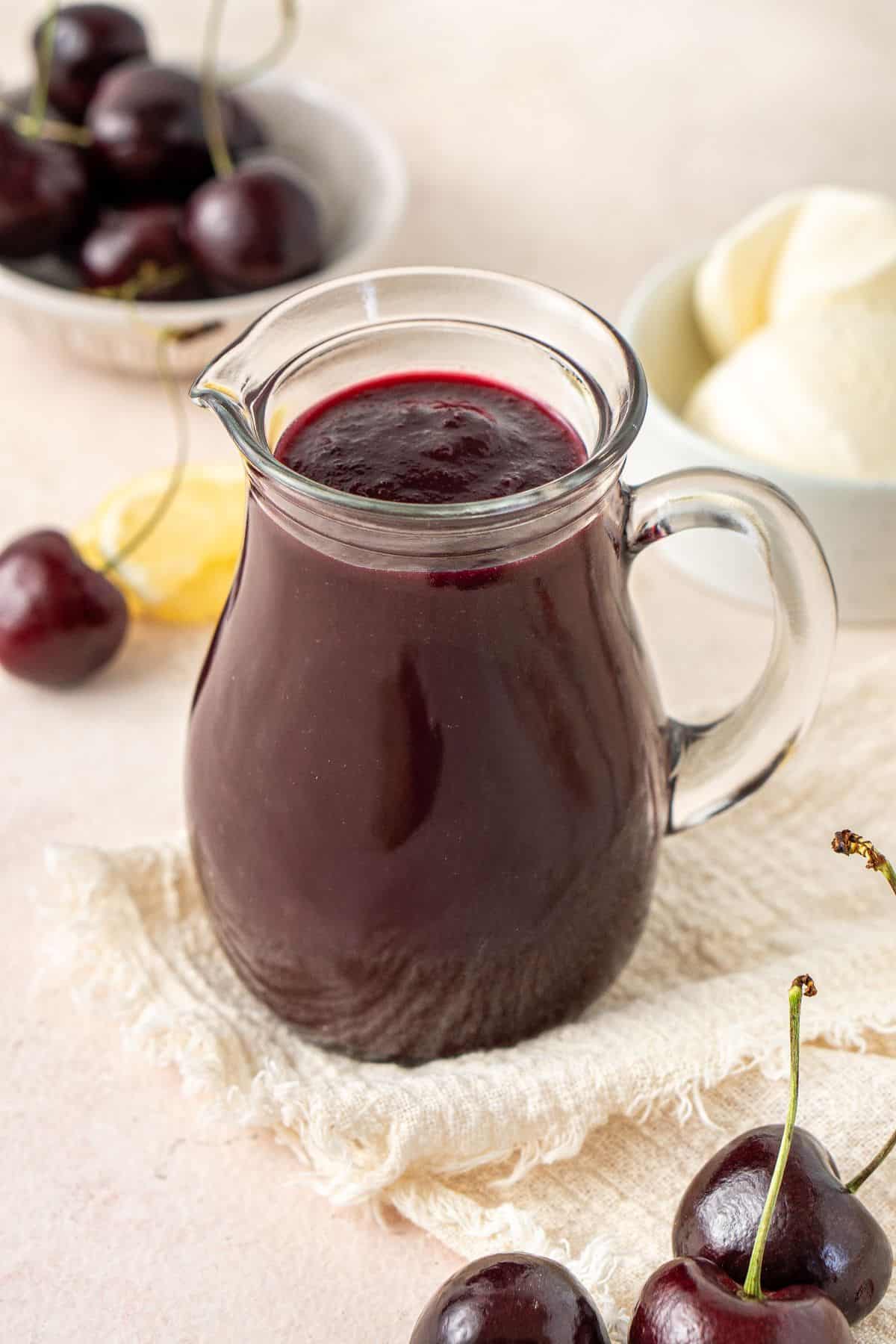Jug of Cherry Sauce, on a cream cloth with some fresh cherries around the edge.