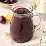 Jug of Cherry Sauce, on a cream cloth with some fresh cherries around the edge.