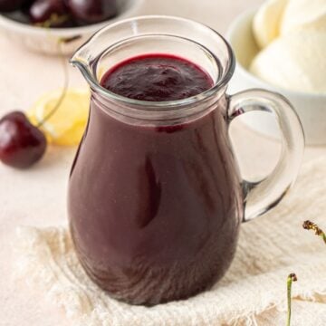 Jug of Cherry Sauce, on a cream cloth with some fresh cherries around the edge.