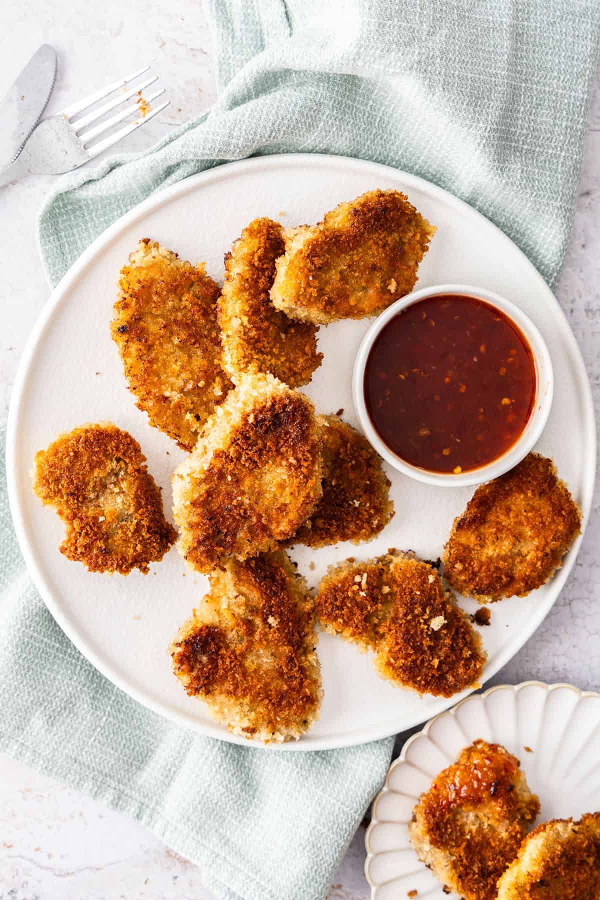 Round white plate with Crumbed Pork Medallions and a small dish of sweet chilli sauce, sitting on a light blue cloth.