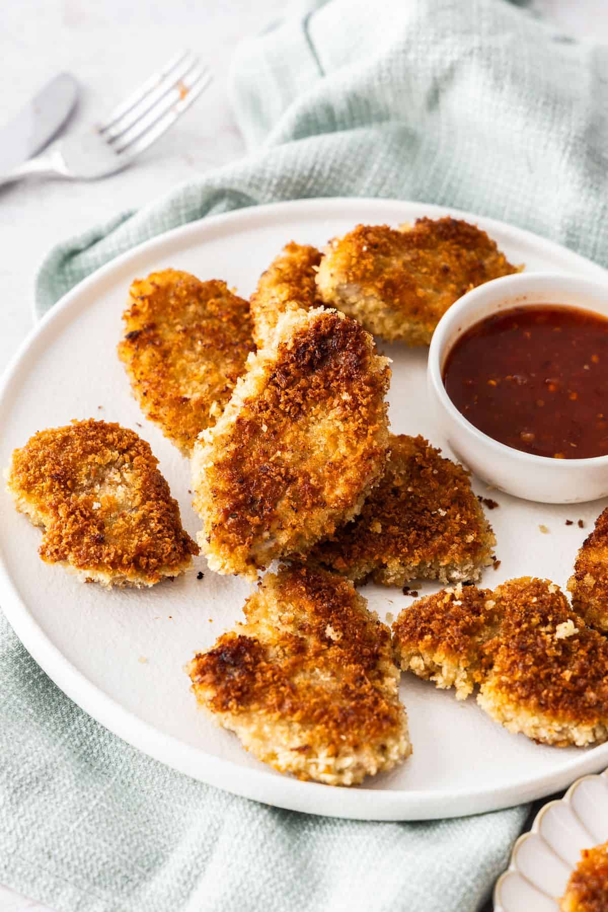 Round white plate with Crumbed Pork Medallions and a small dish of sweet chilli sauce, sitting on a light blue cloth.