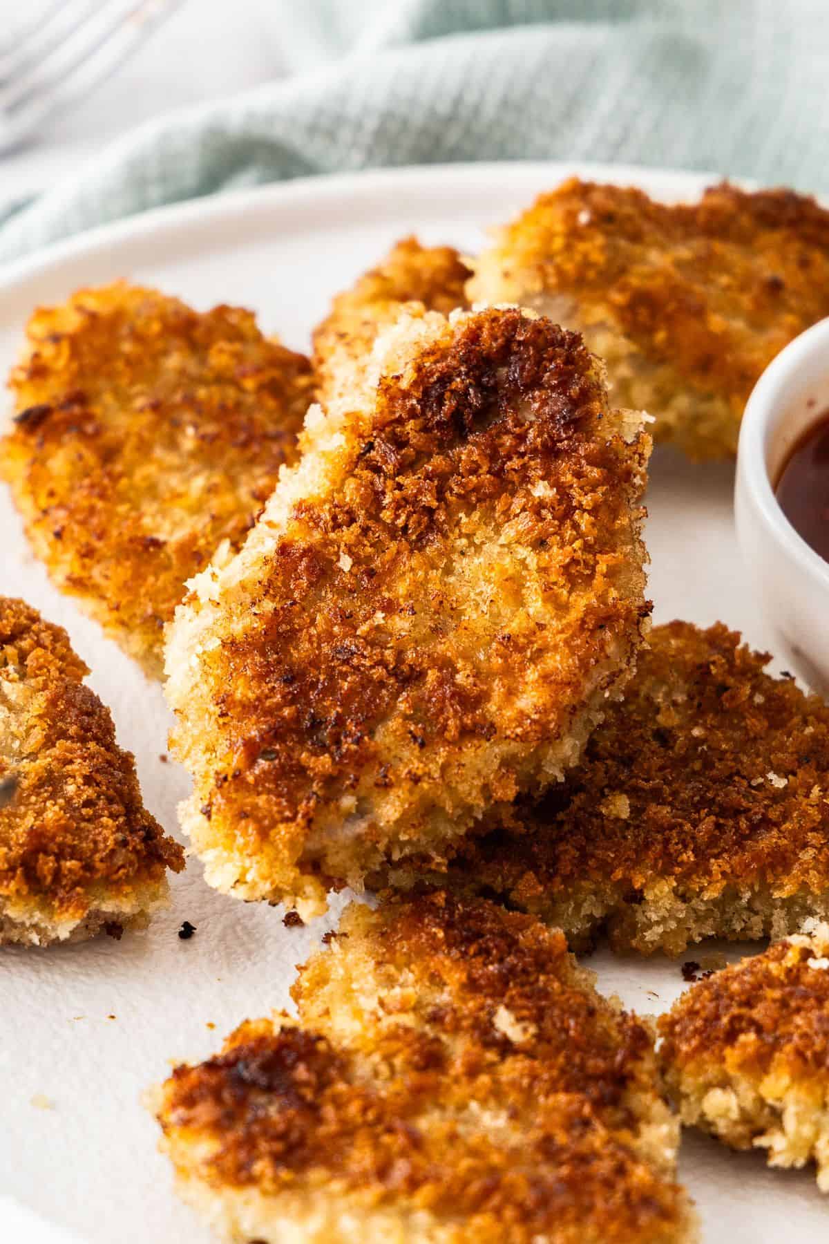 Round white plate with Crumbed Pork Medallions and a small dish of sweet chilli sauce, sitting on a light blue cloth.