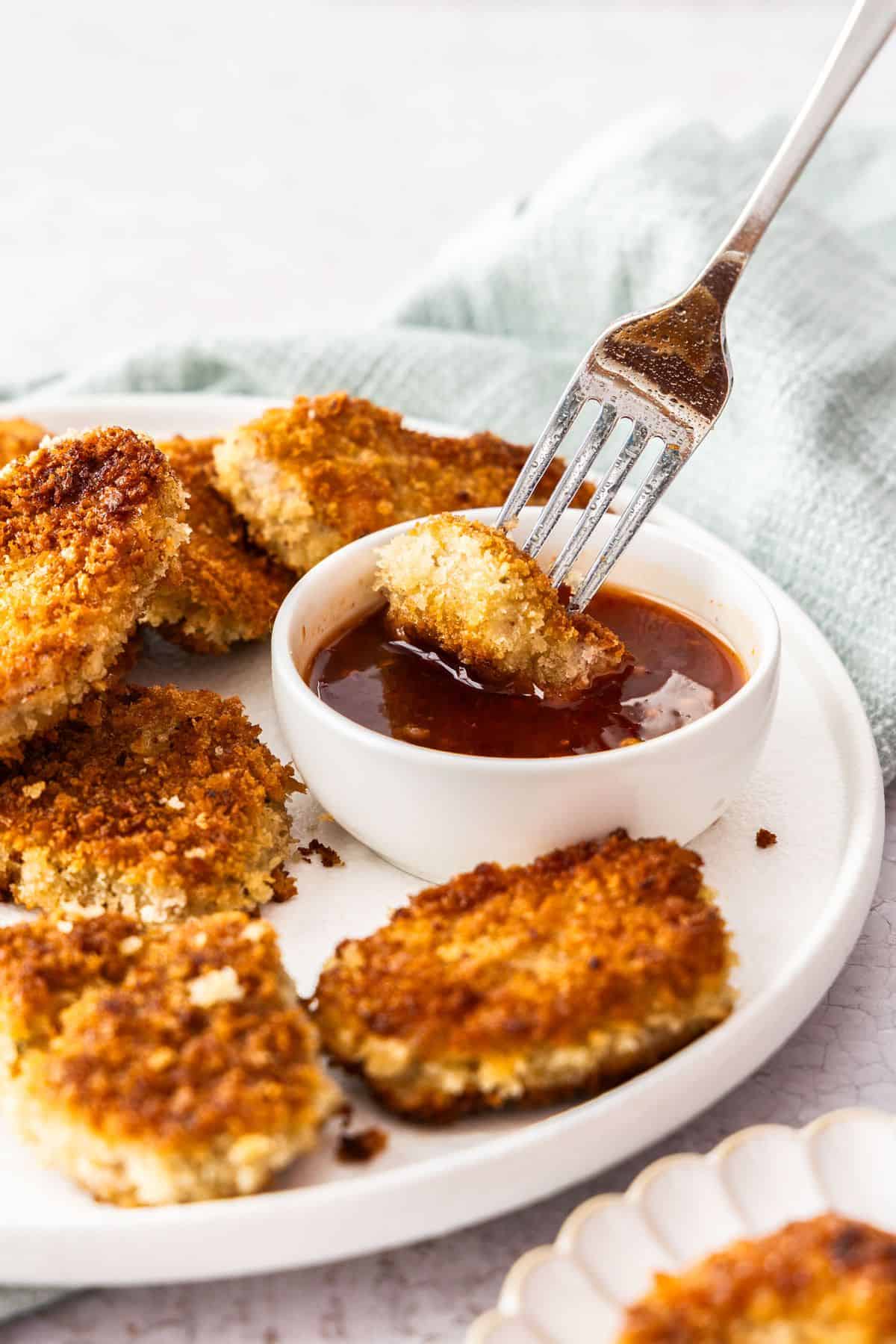 Fork picking up a piece of Crumbed Pork Medallion and dipping it into a small dish of sweet chilli sauce.