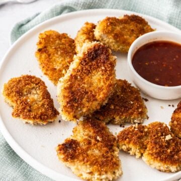 Round white plate with Crumbed Pork Medallions and a small dish of sweet chilli sauce, sitting on a light blue cloth.