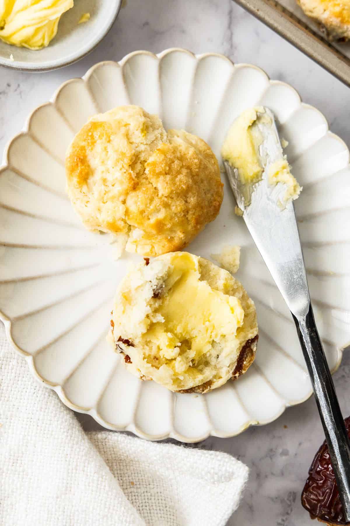 Round plate with a Date and Lemon Scone cut in half, with a knife and some butter on the edge.