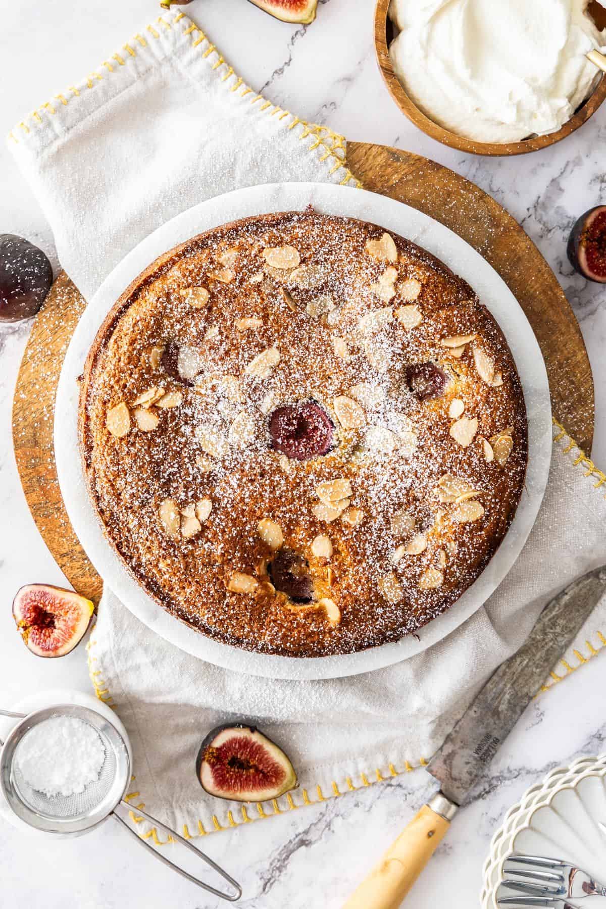 Whole Gluten-Free Fig Cake dusted with icing sugar, on a white plate, sitting on a white cloth with some figs around the edge.