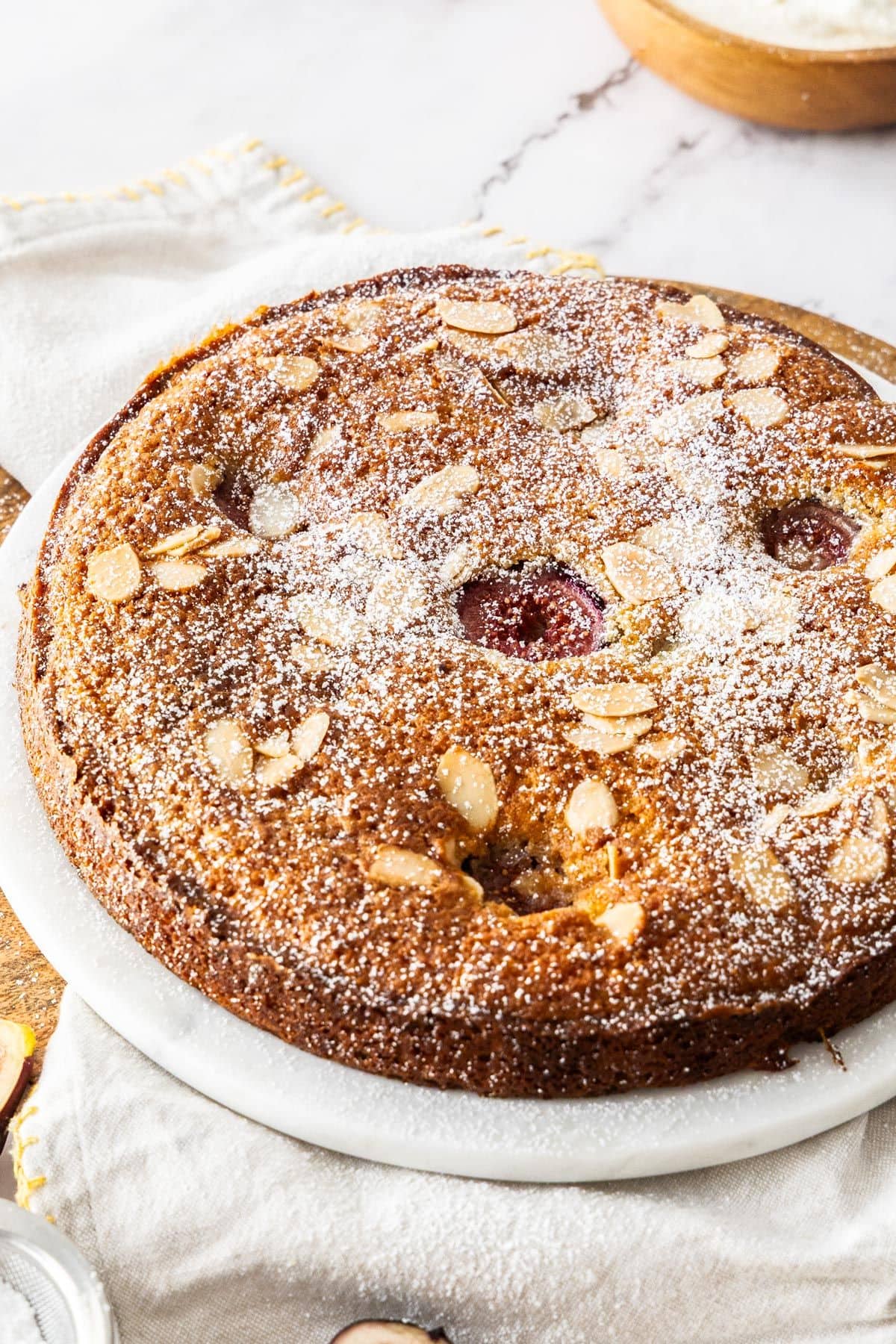 Whole Gluten-Free Fig Cake dusted with icing sugar, on a white plate, sitting on a white cloth.