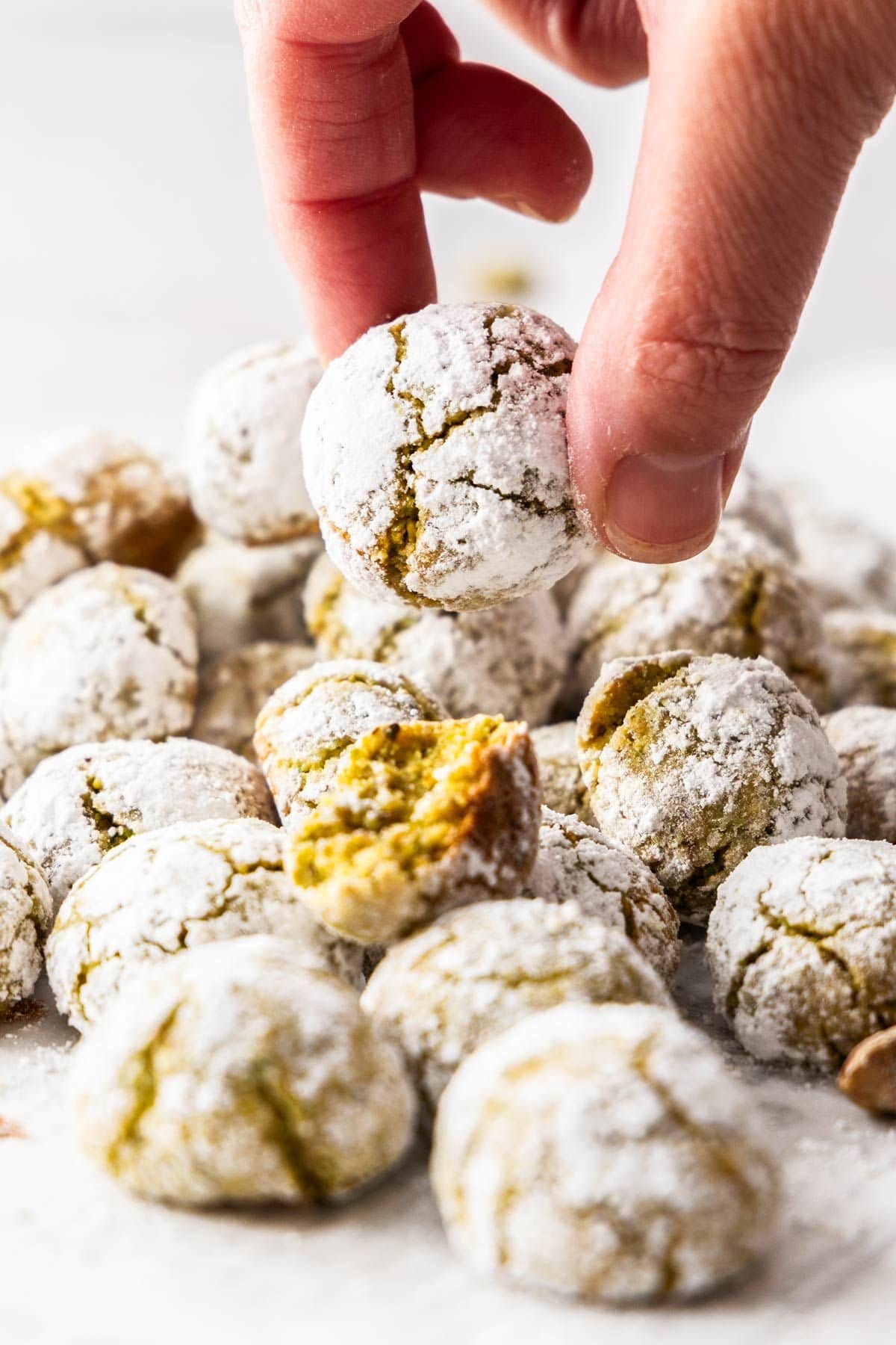 Freshly baked Gluten-Free Pistachio Cookies in a pile on some baking paper, with a hand picking up a cookie.