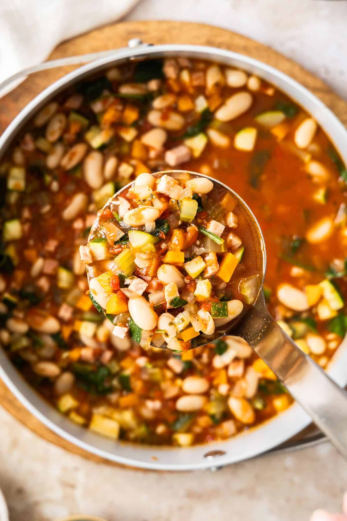Saucepan full of Ham and Cannellini Bean Soup, with a ladle scooping up some soup.