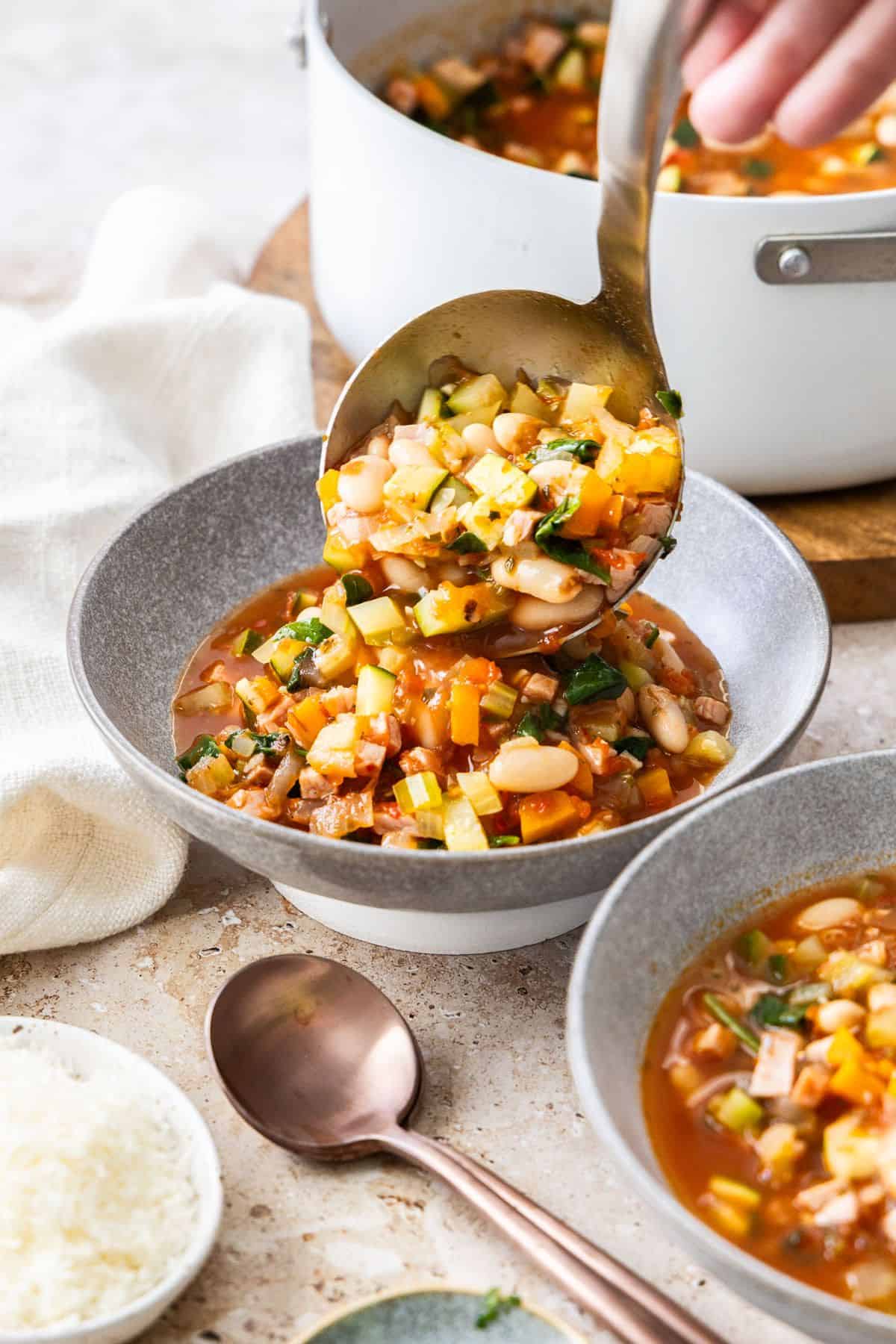 A ladle serving some soup into two small grey bowls, with the saucepan in the background.